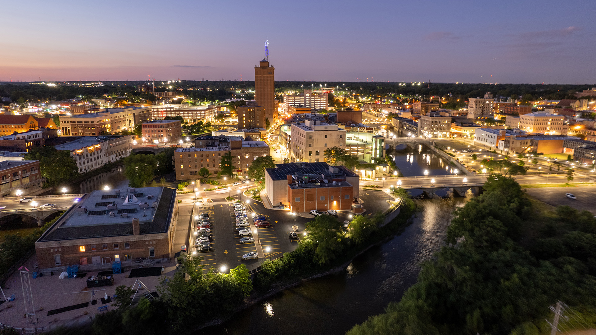 The image is a photograph of an urban night scene featuring a city skyline with illuminated buildings, streetlights, and a prominent clock tower. The foreground shows a river or waterway reflecting the lights, and there are bridges crossing over it. A large parking lot with parked cars is visible on the left side, and a well-lit park area can be seen in the middle ground. The sky is dark, indicating nighttime, and there are no visible texts or distinctive brands in the image.