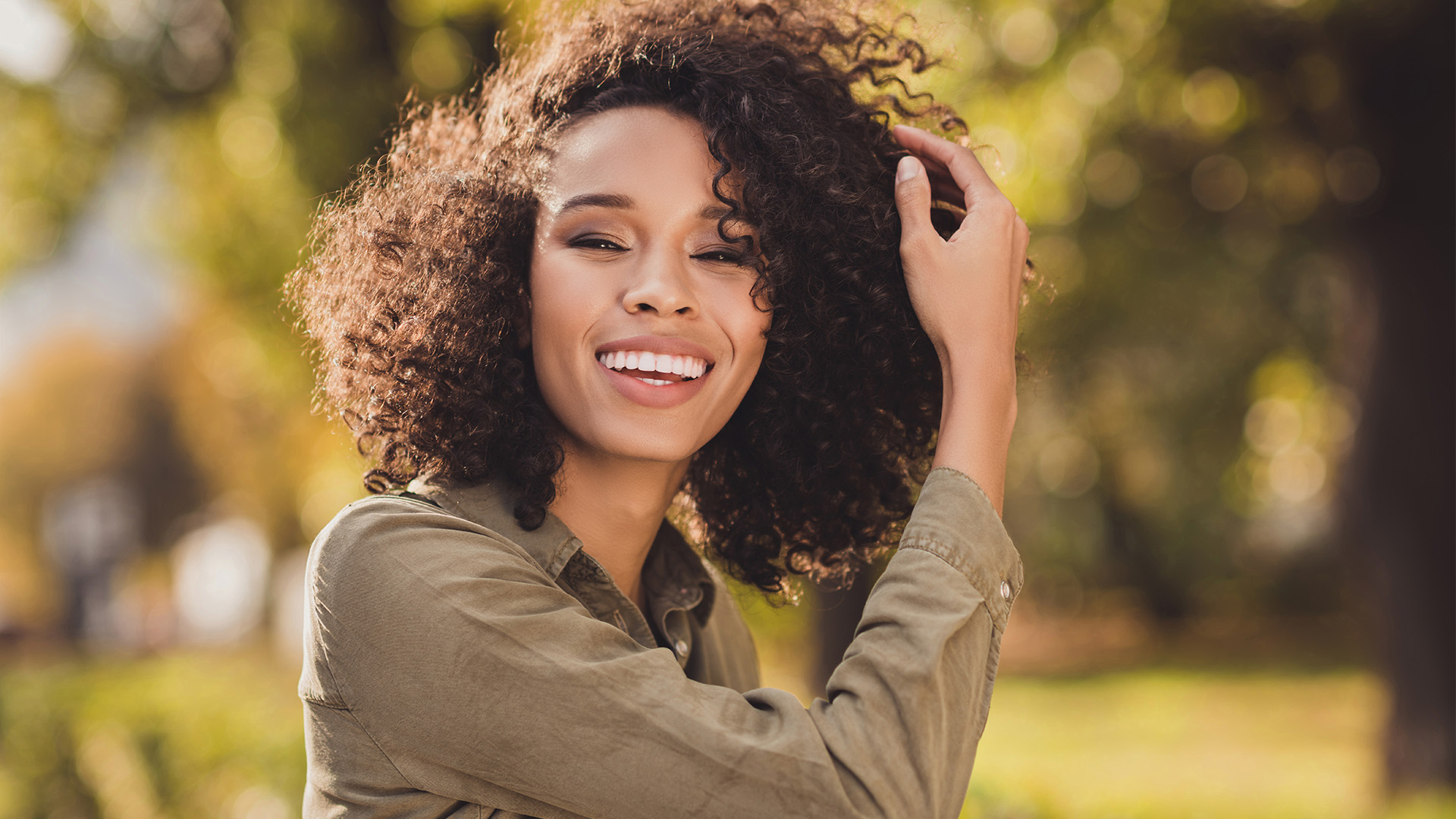 Woman with curly hair smiling, wearing a green jacket and standing outdoors.