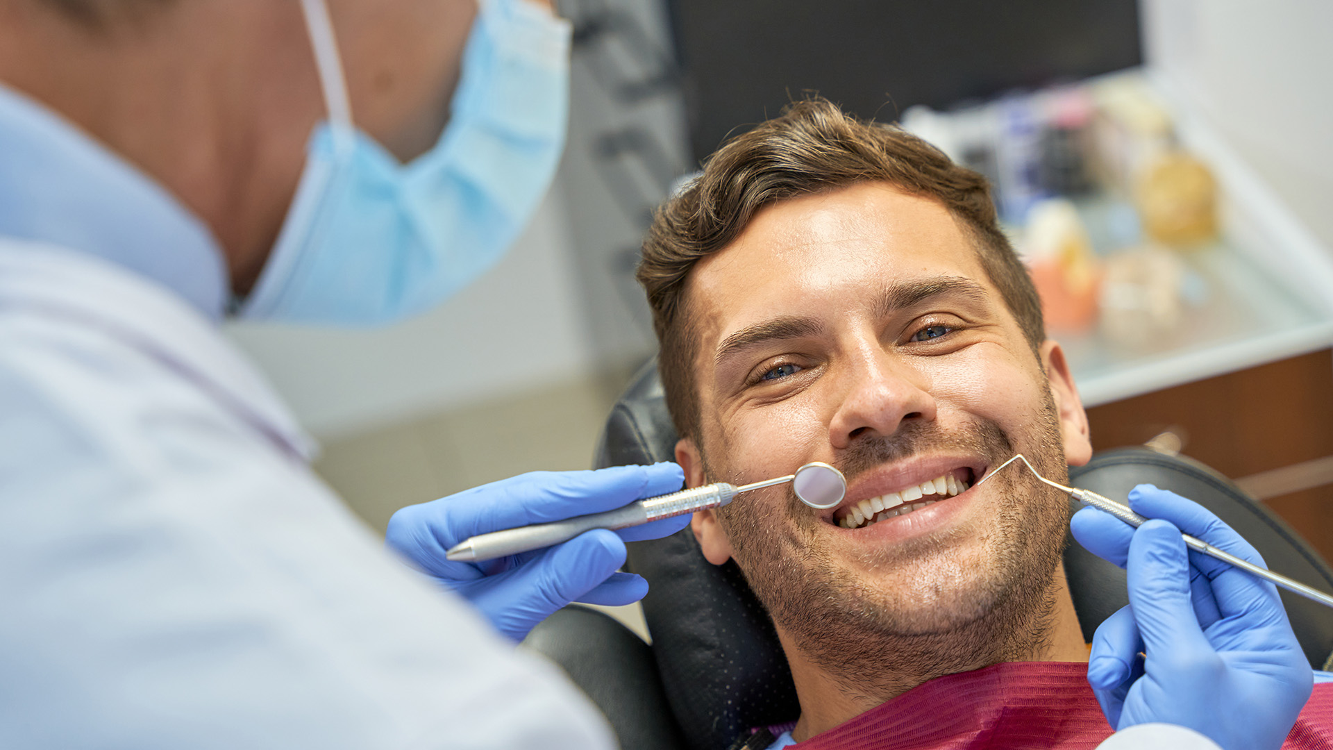 The image features a man sitting in a dental chair, smiling broadly while undergoing a dental procedure. He is wearing an oral appliance and has his eyes closed, appearing relaxed despite the ongoing treatment. A dental professional is attending to him, focused on their work.