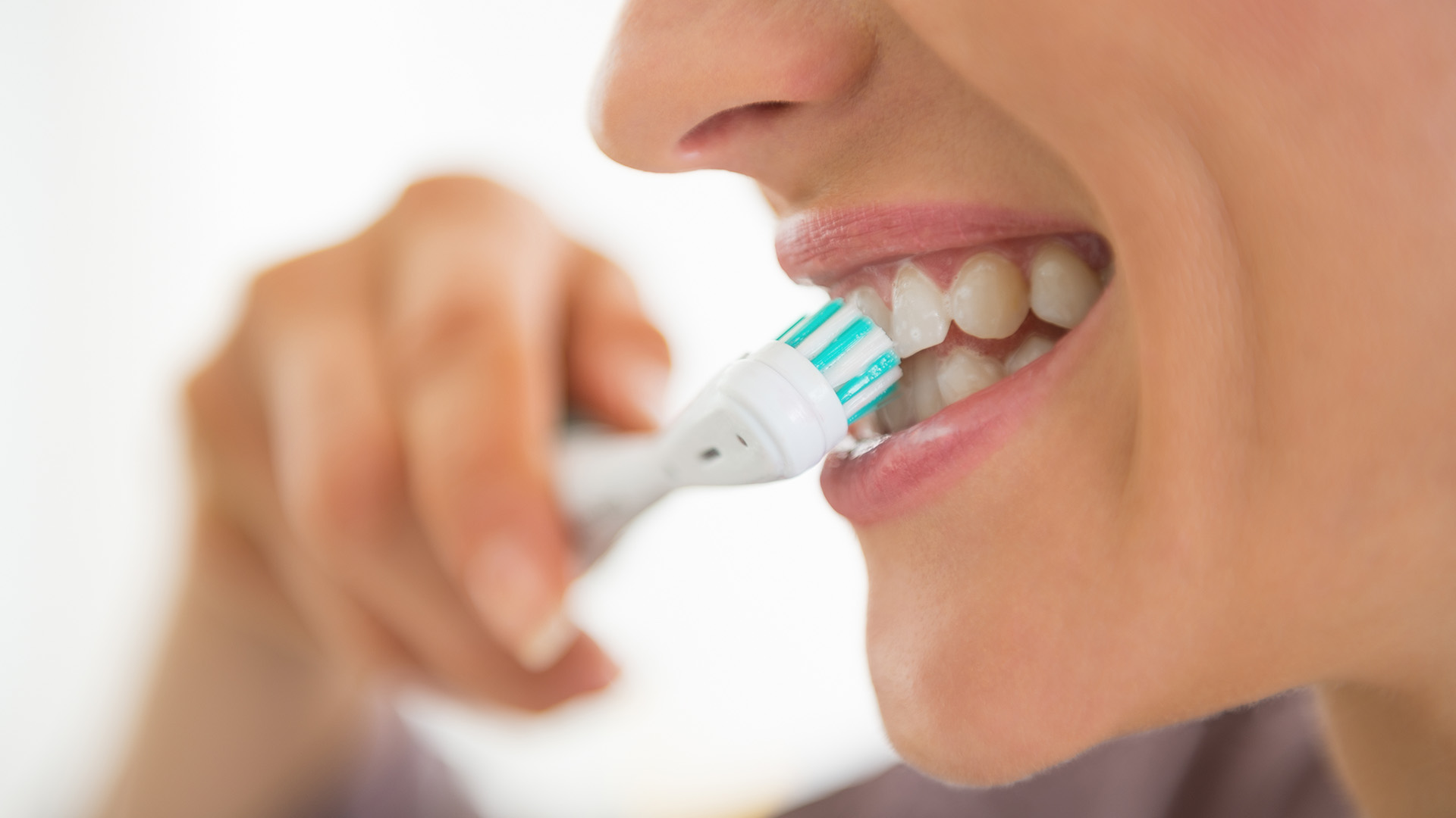 A smiling person brushing their teeth with an electric toothbrush in front of a blurred background.