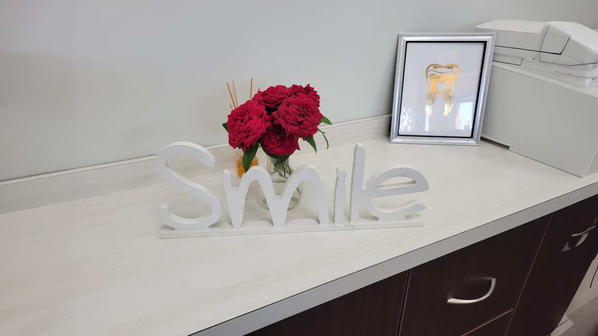 The image shows a smiling sign with the word  smile  on a countertop, placed in front of a framed photo and a potted plant.