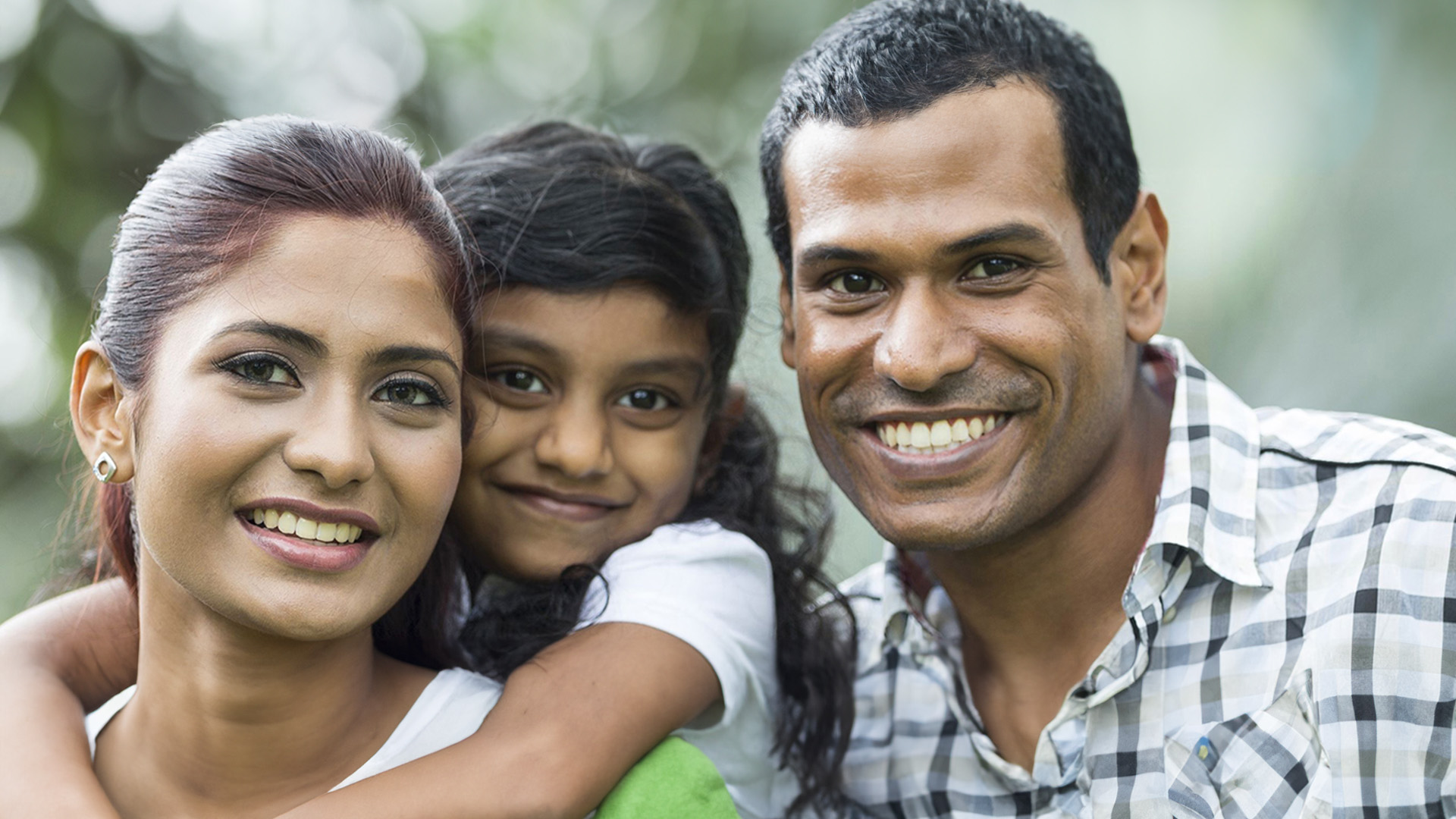 The image is a photograph of three individuals, likely a family, with the father hugging his daughter from behind while smiling. They are outdoors during daylight.