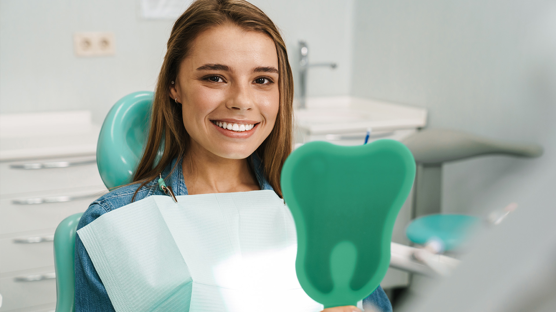 A young woman is sitting in a dental chair, smiling at the camera with a mouthguard on her teeth.