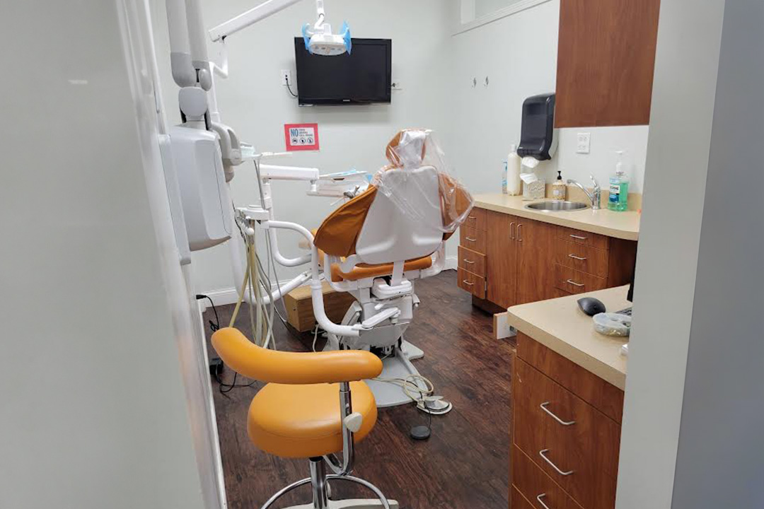 The image shows an interior view of a dental office, featuring a dental chair, equipment, and a workstation with a computer monitor.