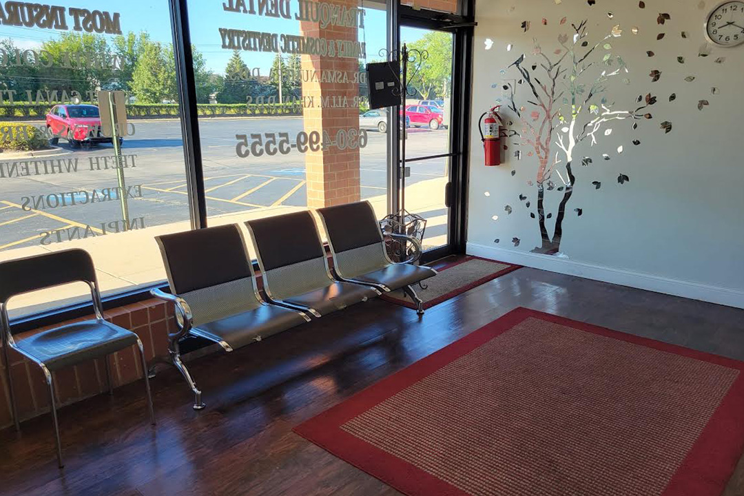 The image shows an interior space, likely a waiting area or lobby, with a red carpet on the floor leading to a set of double doors. There are two benches facing each other, and a small seating area with chairs is visible in the background. A wall-mounted sign with decorative elements is present above the benches. The room has a modern design with neutral colors and natural light from a window on the left side of the image.