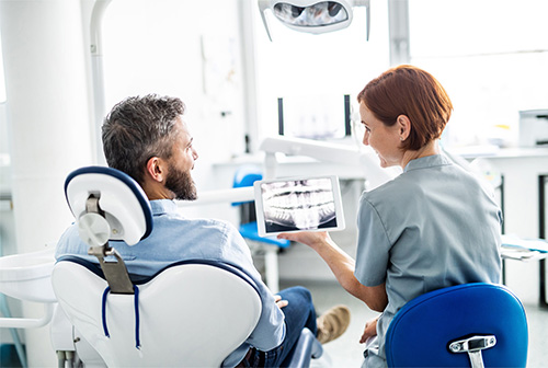 The image depicts a dental office setting where a dentist is seated in a chair, holding an iPad displaying a patient s digital dental records, while a dental hygienist stands beside him, observing the screen.