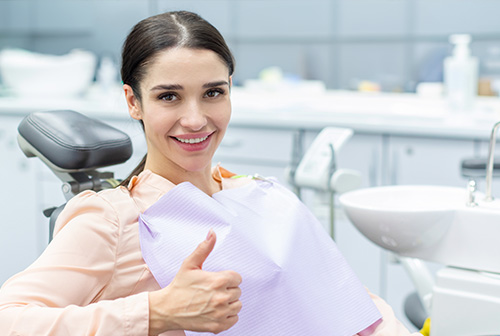 The image shows a woman sitting in a dental chair, smiling and giving a thumbs-up gesture. She is wearing a pink top and appears to be in a dentist s office setting, with dental equipment visible in the background.