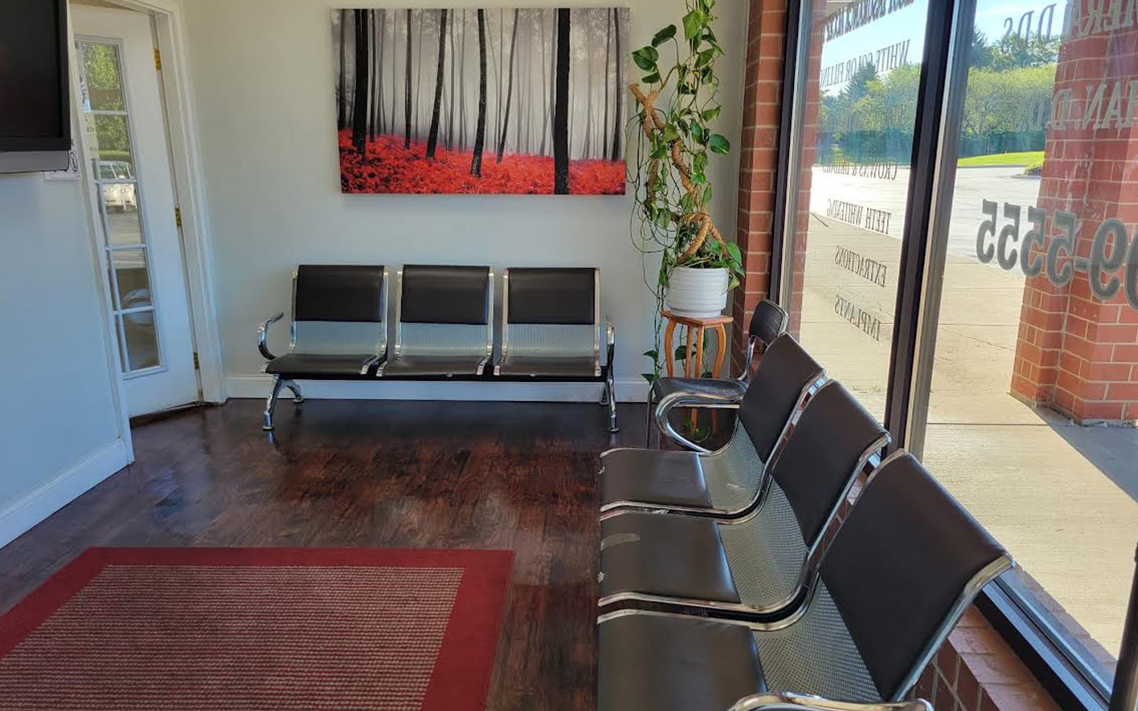 The image shows an interior space, likely a waiting area or lobby, with a row of black chairs facing each other, a red carpet on the floor, and a large window that allows natural light to enter.