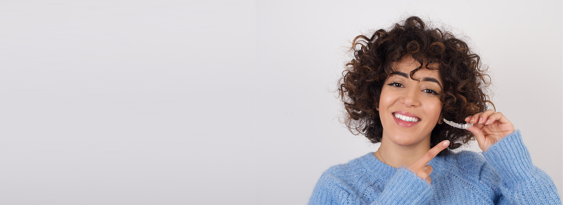 A smiling woman with curly hair, wearing a blue sweater, stands against a white background.