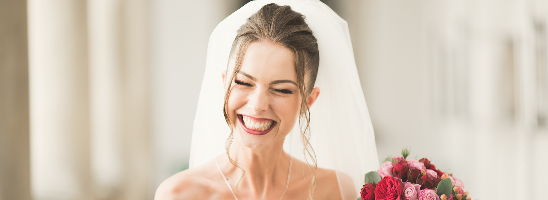 The image is a photograph of a smiling woman wearing a white bridal veil, standing outdoors with her hair up, and in the foreground, there s a watermark or logo overlaying the image.