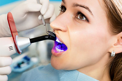 The image shows a woman seated in a dental chair, receiving a dental cleaning with a device that sprays water or air, while a dental hygienist uses a handheld tool to clean her teeth.