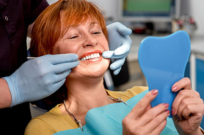 The image shows a woman in a dental chair, smiling broadly while holding a blue dental impression between her fingers. She is being attended to by a dentist who is adjusting the impression with gloved hands.