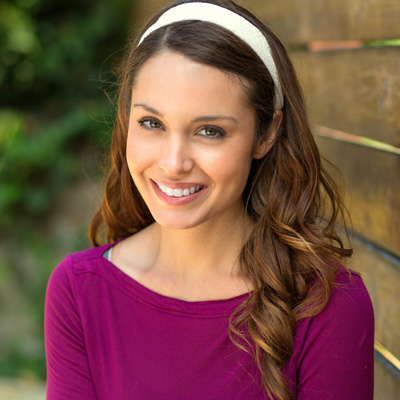 A woman with long brown hair is smiling, wearing a purple top and a headband, standing in front of a wooden fence.