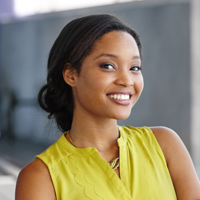 A smiling woman with a yellow top and a hair tie, posing against a blurred background.
