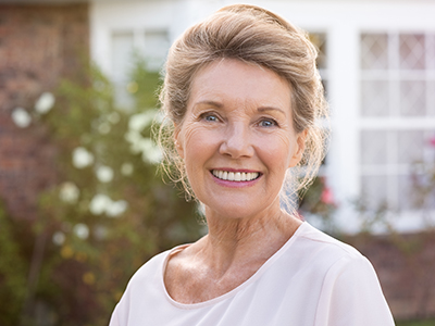 An elderly woman with blonde hair, wearing a white top and smiling at the camera, standing in front of a brick house with a garden.