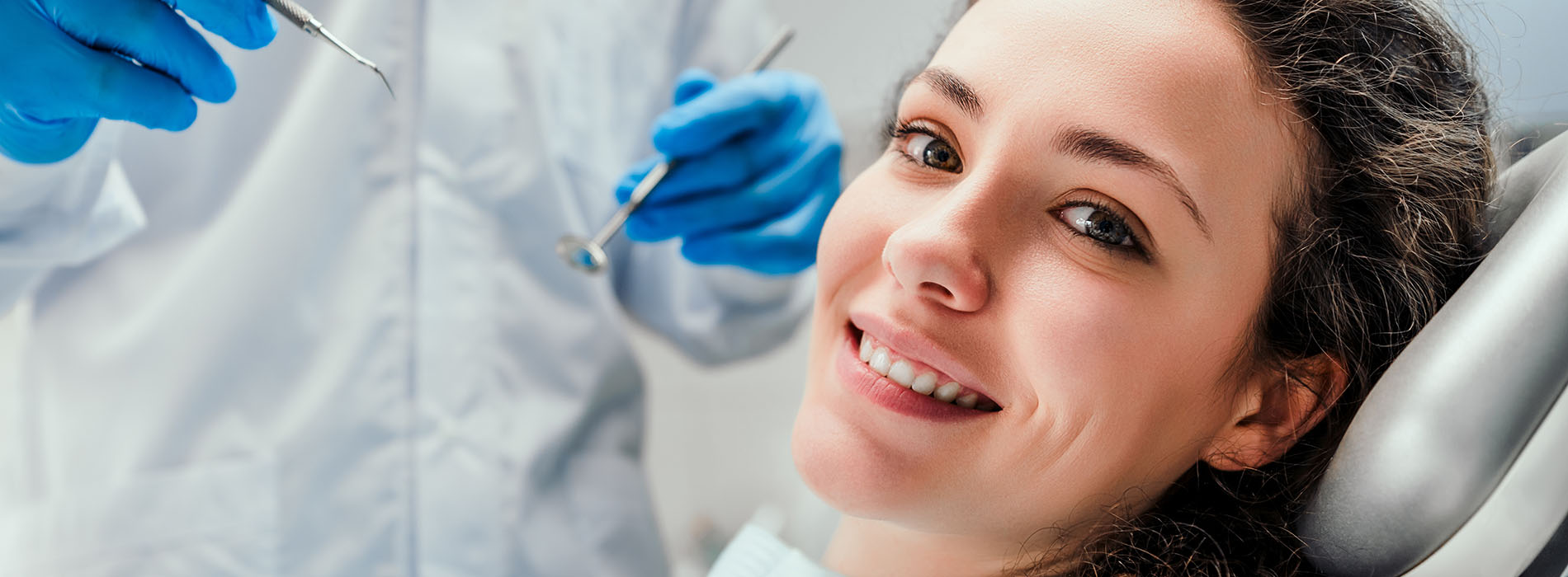 A woman is seated in a dental chair, smiling at the camera, while a dental professional attends to her.