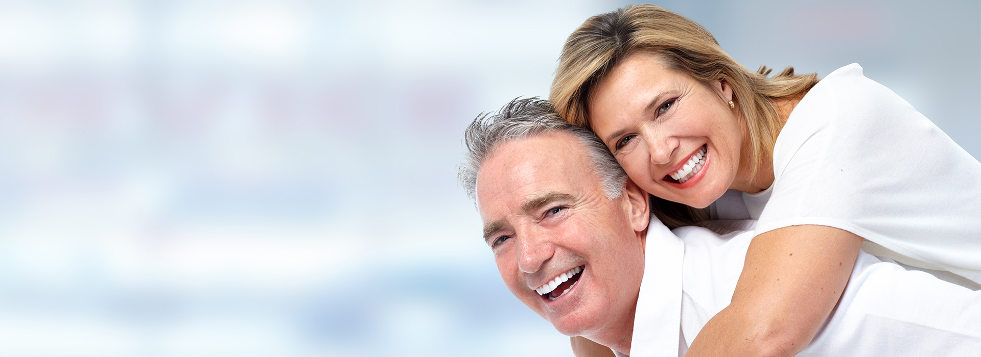 The image depicts a man and woman smiling at the camera, with the man wearing glasses and both appearing to be in their senior years. They are embracing each other in a joyful manner.