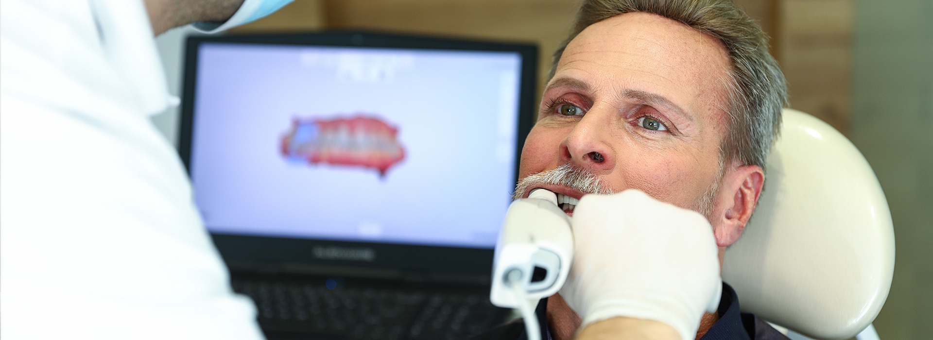 The image shows a dental professional performing a dental procedure, with the patient seated in an examination chair and the professional using a computer monitor displaying what appears to be an X-ray or digital imaging of the patient s mouth.