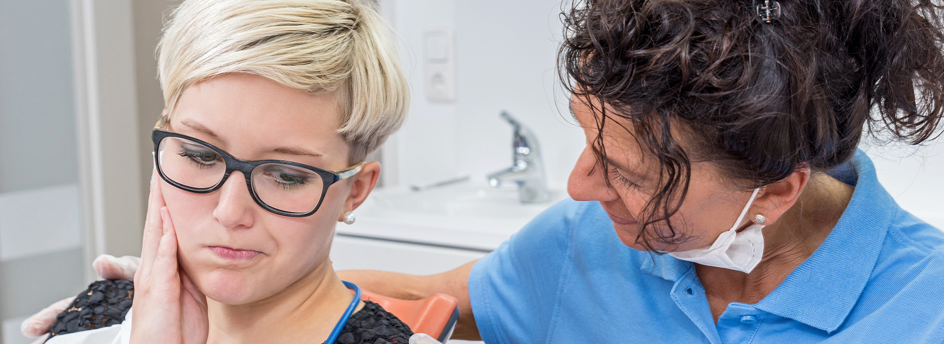 A woman with short hair is sitting in a dental chair, receiving dental care from a professional wearing blue glasses and a blue shirt.