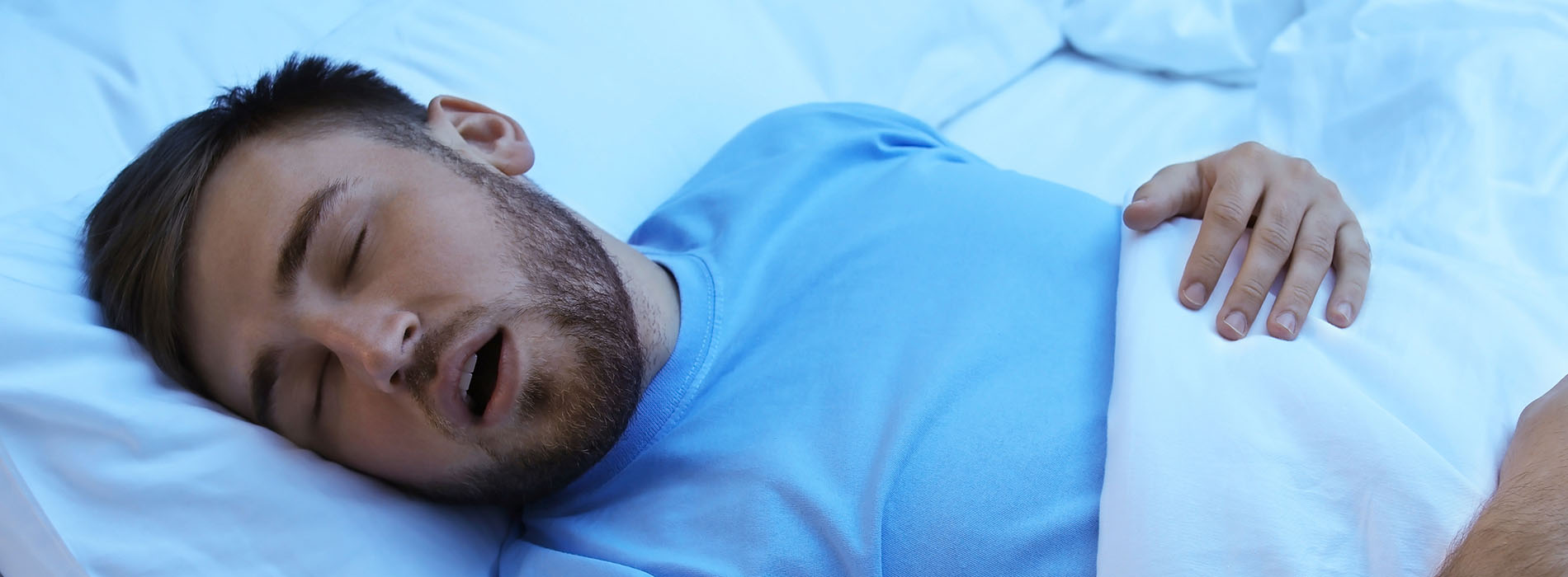 A man is lying in bed with his mouth open, appearing to be asleep or possibly in the middle of a sleep-talking episode.