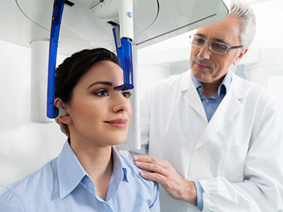 A medical professional stands next to a woman receiving a 3D facial scan, with the machine displaying her face in 3D.