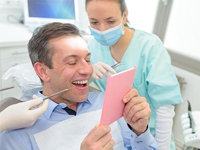 In the image, a man is seated in a dental chair, holding up a pink card with a surprised expression. A woman wearing a surgical mask stands behind him, holding a tray of dental instruments. The man appears to be at a dentist s office, and there is a computer screen displaying an image of a smiling face in the background.