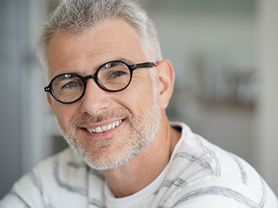 The image shows a man with short hair, wearing glasses and a beard, smiling at the camera. He has graying hair and is dressed in casual attire.