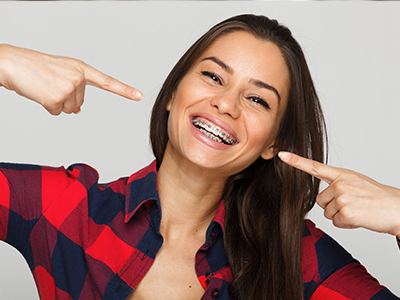 A woman with a wide smile pointing at her teeth, wearing a plaid shirt and standing against a white background.