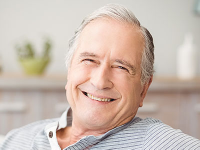 The image shows a smiling older man with gray hair, wearing a blue shirt and seated in a relaxed position.