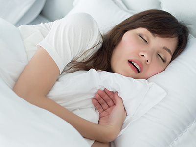 The image depicts a woman lying in bed with her eyes closed, wearing a white top and holding a pillow. She appears to be asleep or resting with her head on the pillow.