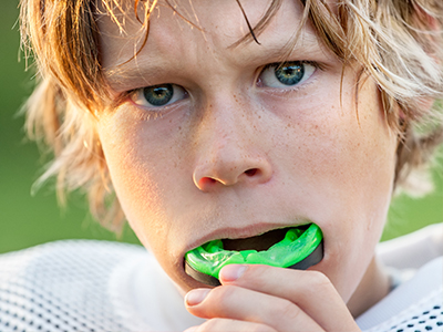 A young boy with blonde hair and a determined expression is holding a green toothbrush in his mouth, which he has just bitten.