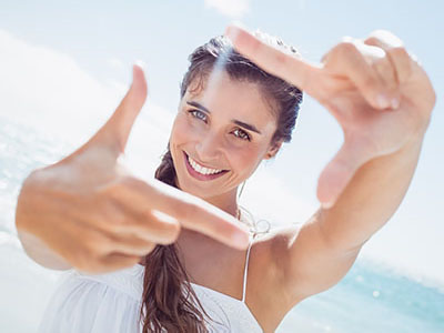 The image shows a smiling woman taking a selfie with her right hand, while her left hand is raised to frame the shot. She has long hair and is wearing a white top. The background suggests a beach setting with clear skies.