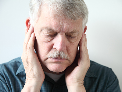 The image is a photograph of an older man with his eyes closed, holding his head as if in pain or discomfort. He has gray hair and appears to be indoors with a plain background.